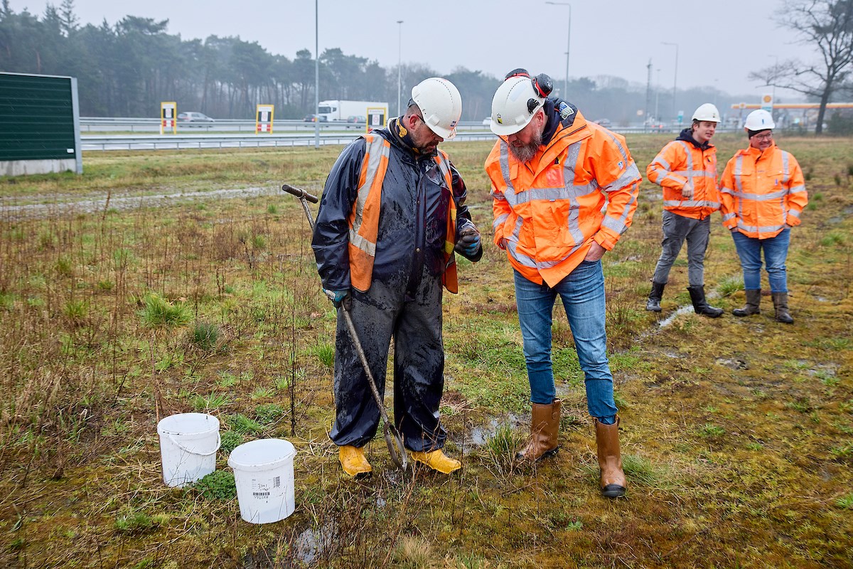 Vier mensen in oranje werkkleding nemen grondmonsters in veld langs de snelweg