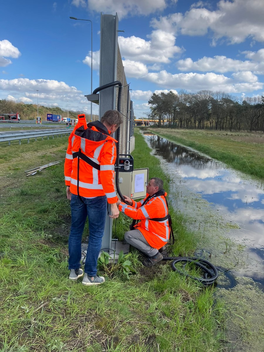 Twee mensen in oranjewerkkleding werken aan irrigatiesysteem aan mosbeton geluidscherm langs de snelweg