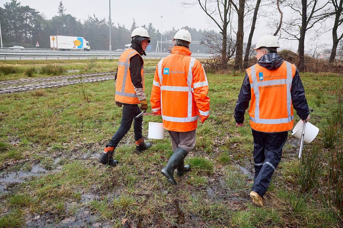 Drie mensen in oranje werkkleding lopen door weiland langs de snelweg